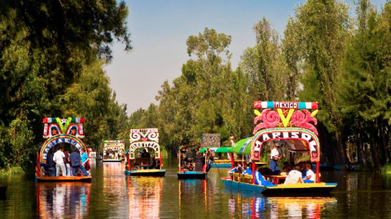 Recorrido en Trajinera Gay en Xochimilco Trajineras coloridas en un canal rodeado de árboles, con casas flotantes decoradas.